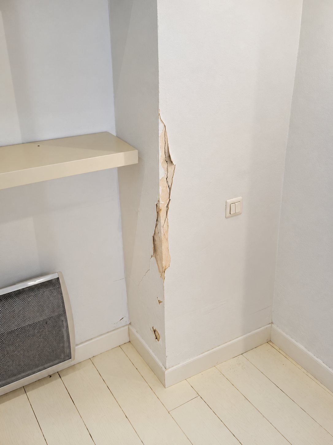 Angle d'un mur blanc dont la peinture s'écaille et se fissure, à côté d'une étagère, d'un radiateur gris et d'un parquet blanc.