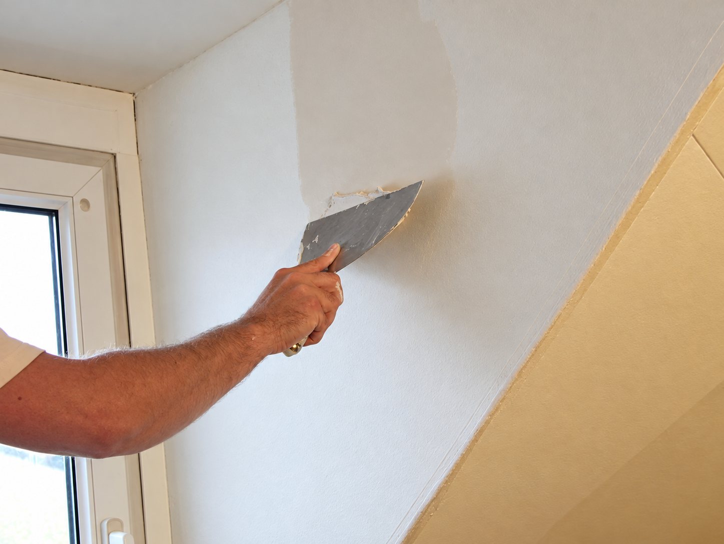 Une main tient un couteau à enduire et lisse un mur blanc dans une pièce sous les combles, près d'une fenêtre.