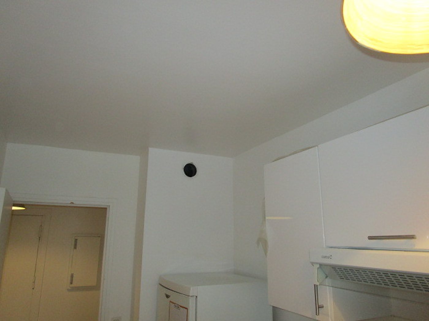 Intérieur d'une petite cuisine aux murs et au plafond blancs, avec meubles hauts laqués, hotte, lave-linge et bouche d'aération noire.