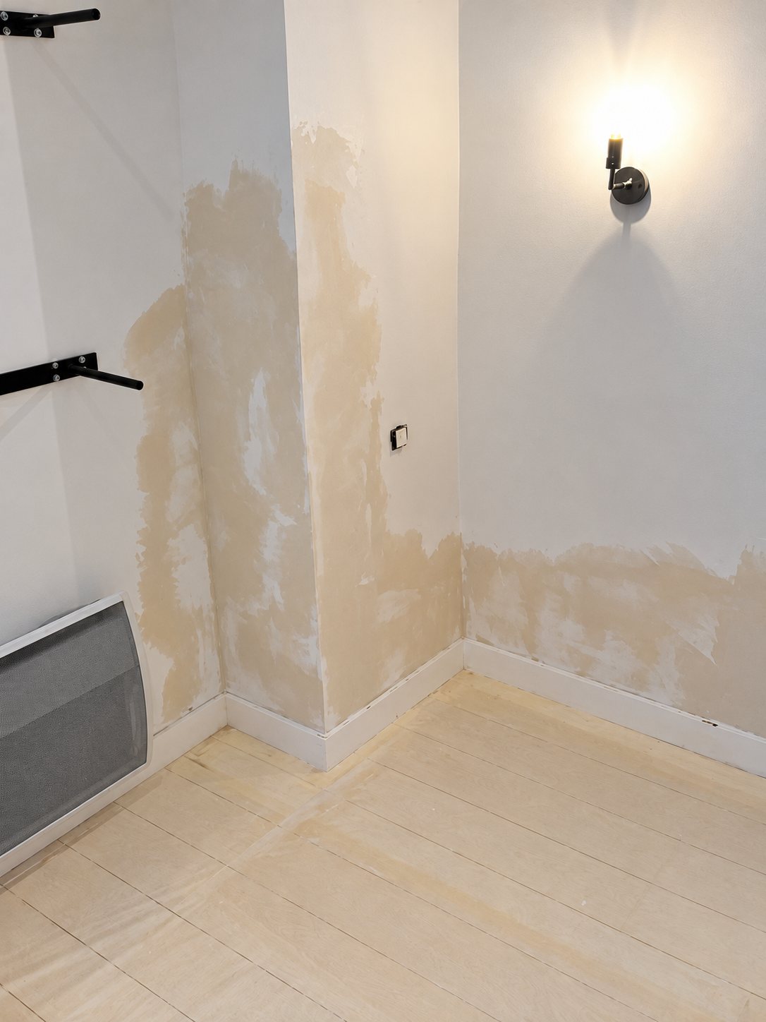 Angle d'une pièce vide dont les murs blancs présentent de larges taches d'humidité beige, avec un radiateur gris et une applique allumée.