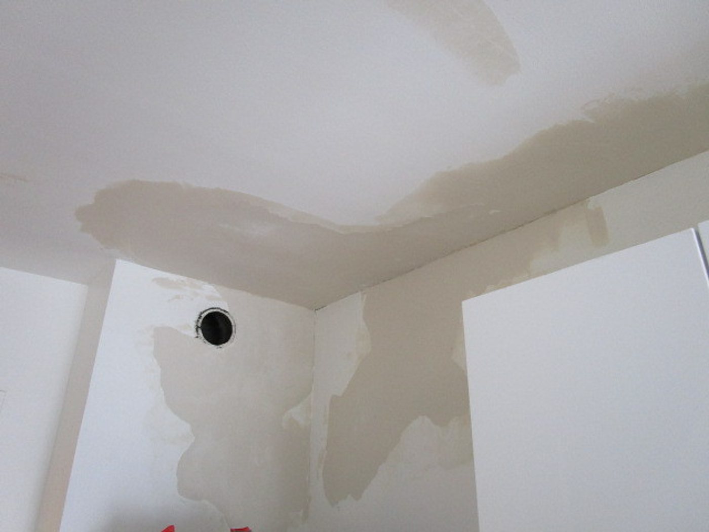 Angle d'une pièce où le plafond et le mur blancs présentent de larges taches beiges d'humidité et de reprises d'enduit, avec une petite ouverture ronde noire dans le mur.