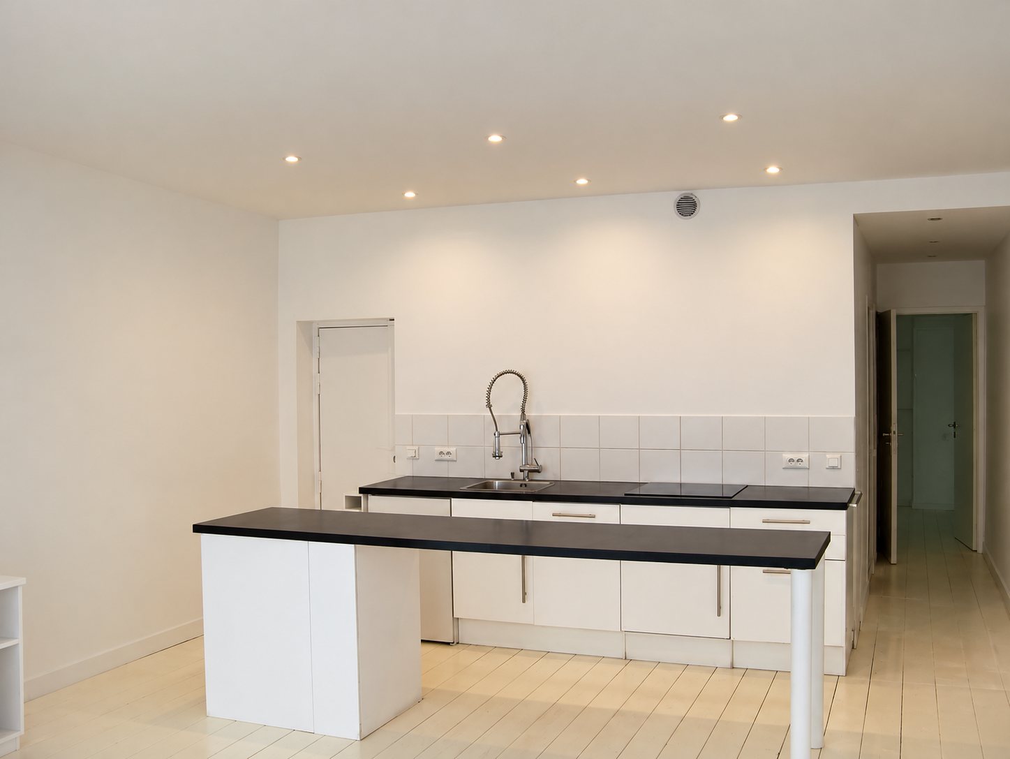 Une cuisine moderne aux meubles blancs et plans de travail noirs, avec îlot central et spots au plafond, dans un appartement rénové au parquet blanc.