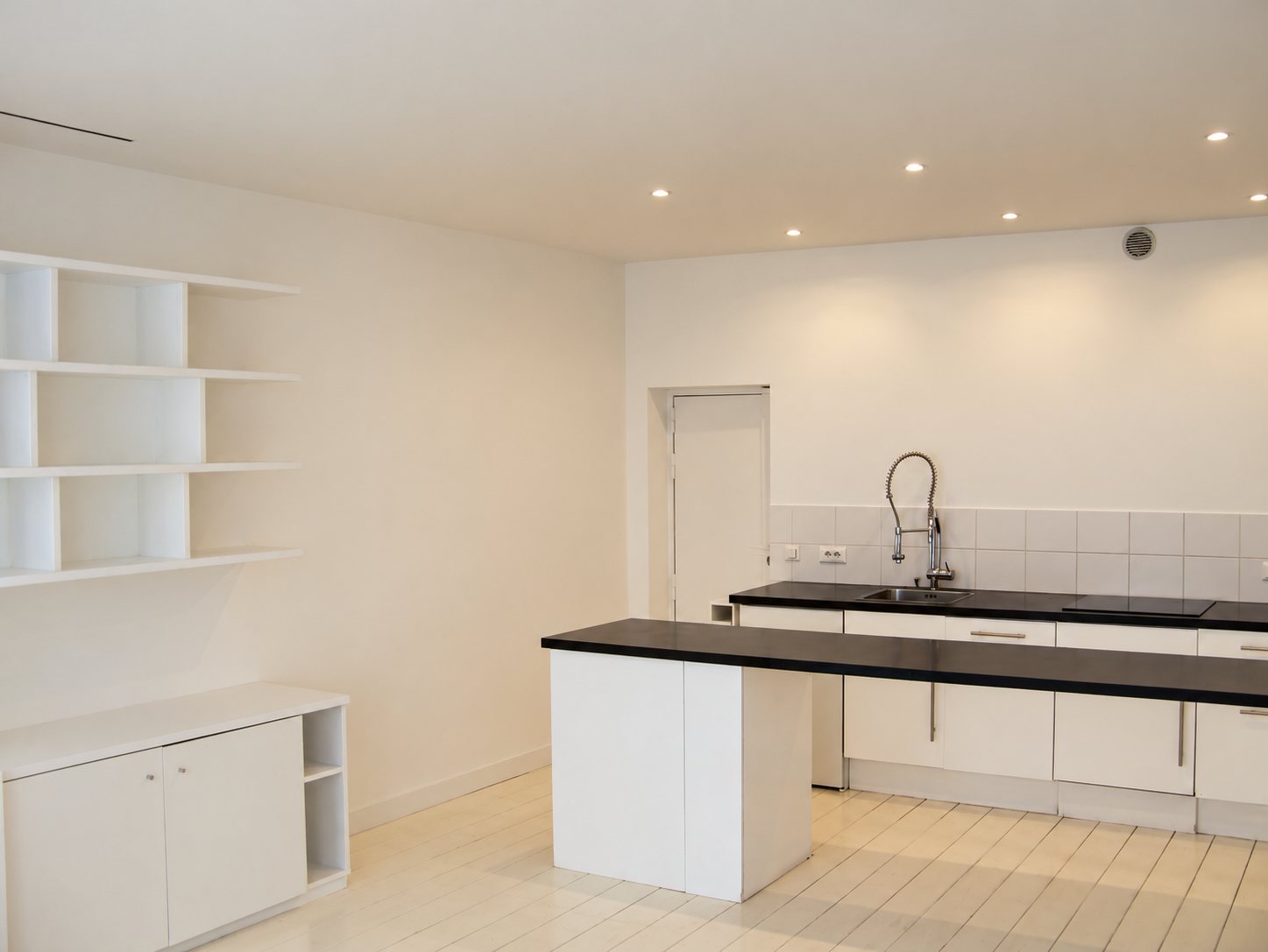 Une cuisine moderne aux murs blancs avec un îlot central, des plans de travail noirs, une robinetterie de type professionnel et des étagères ouvertes sur la gauche.