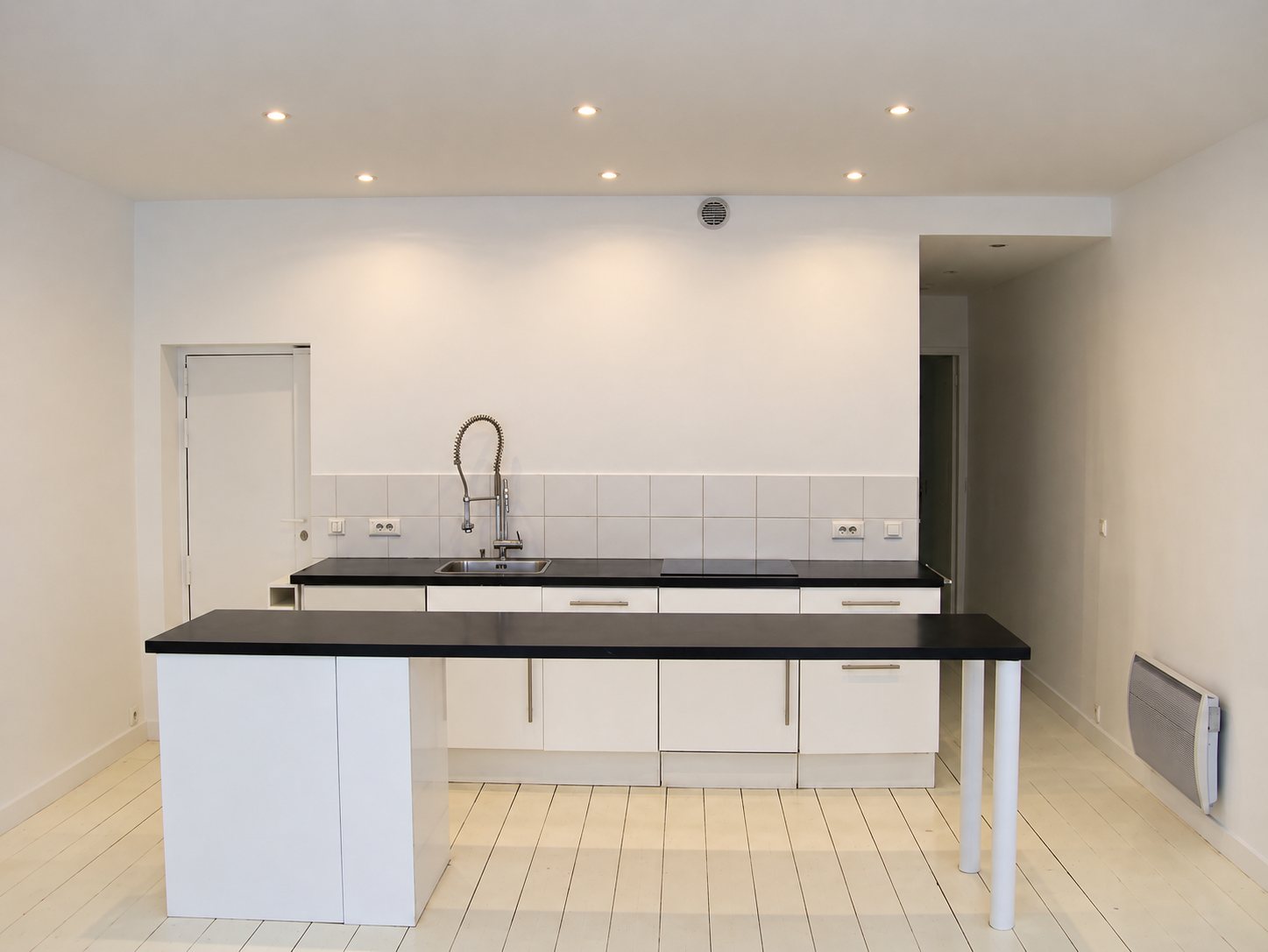 Cuisine ouverte rénovée aux meubles blancs et plans de travail noirs, avec îlot-bar, évier à douchette et parquet peint en blanc.