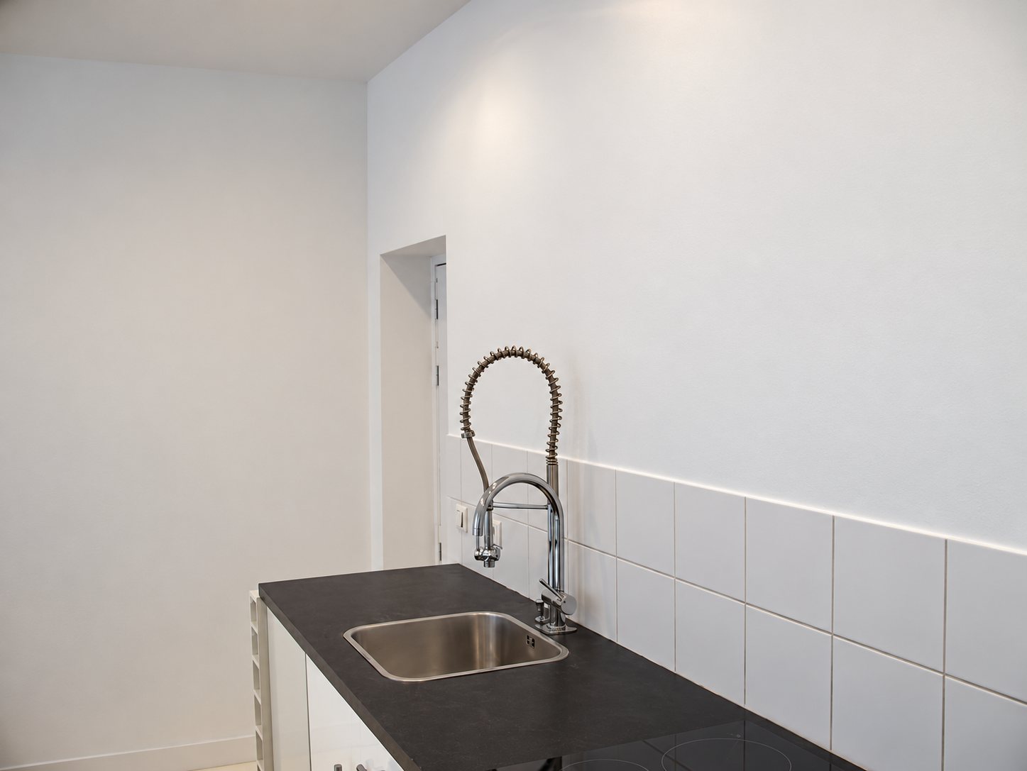 Plan de travail sombre avec évier inox et robinet à ressort, devant un mur blanc et une crédence en carreaux blancs dans une cuisine rénovée.