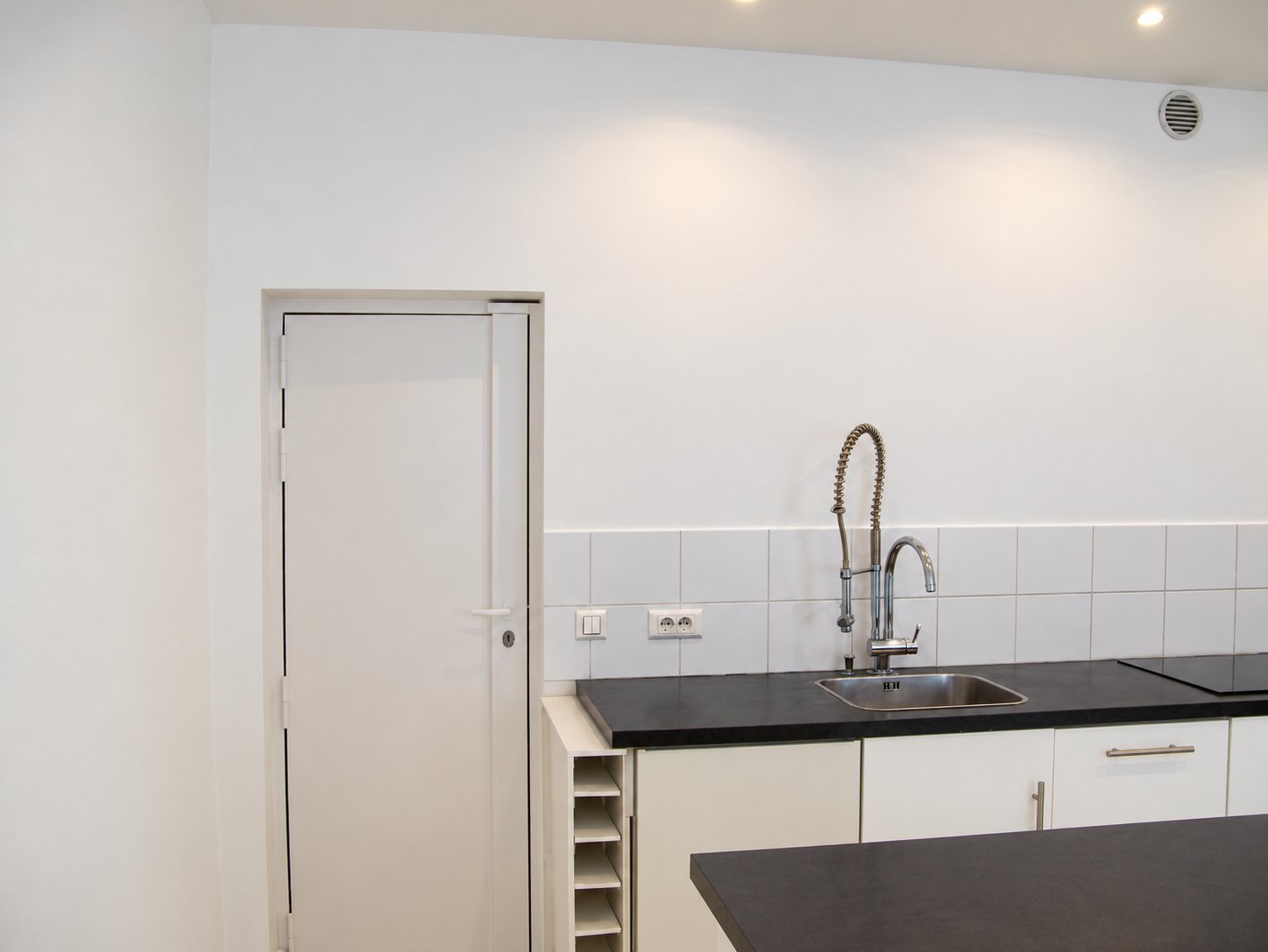 Cuisine moderne fraîchement rénovée avec murs blancs, porte blanche, robinet douchette en inox, plan de travail noir et îlot central.