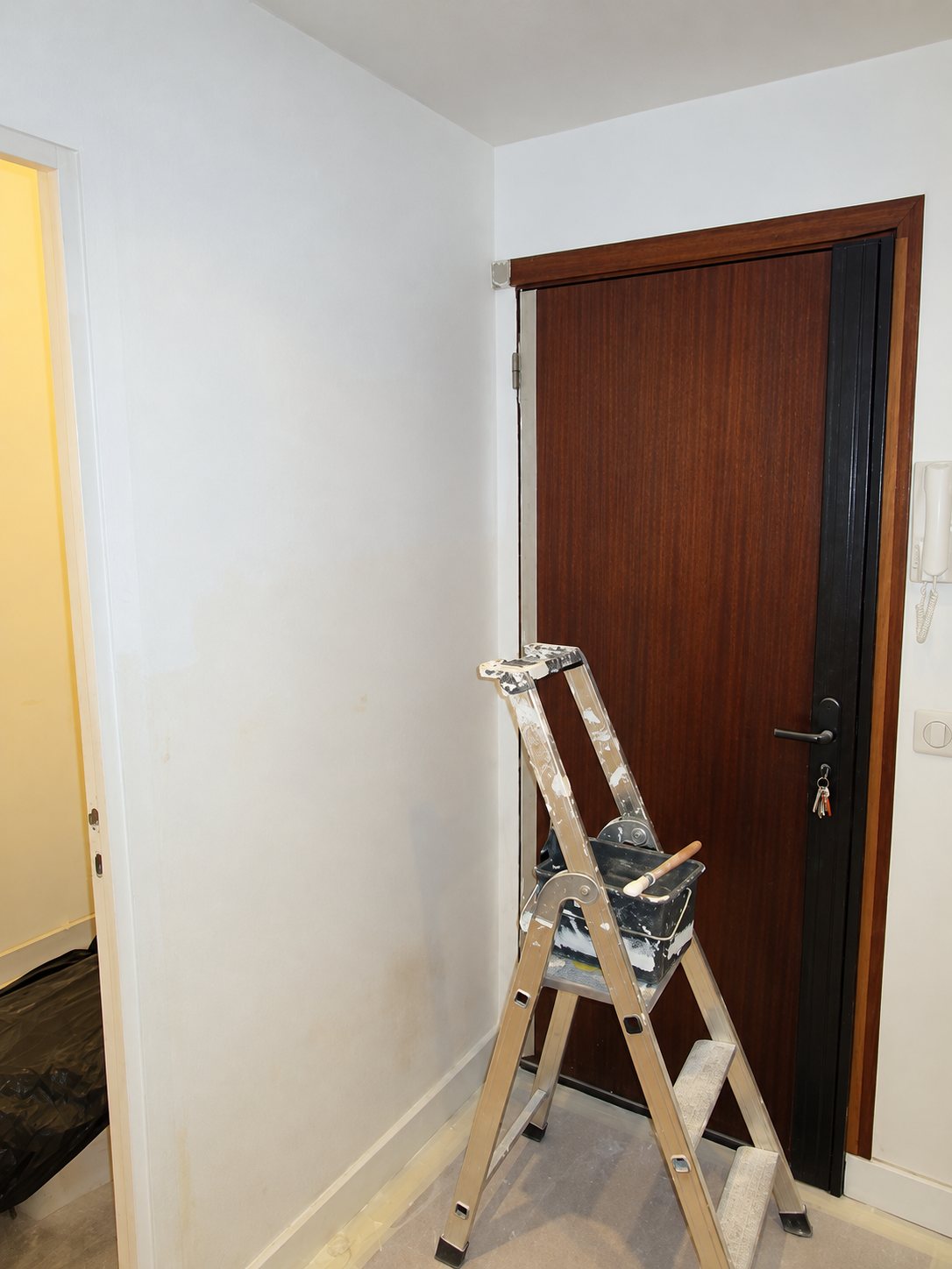 Un escabeau avec un bac de peinture et un pinceau est posé dans une entrée aux murs blancs fraîchement peints, devant une porte en bois foncé.
