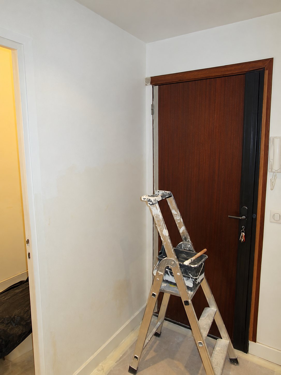 Un escabeau taché de peinture avec un pot et un pinceau se tient dans un couloir aux murs blancs, devant une porte d'entrée en bois foncé.