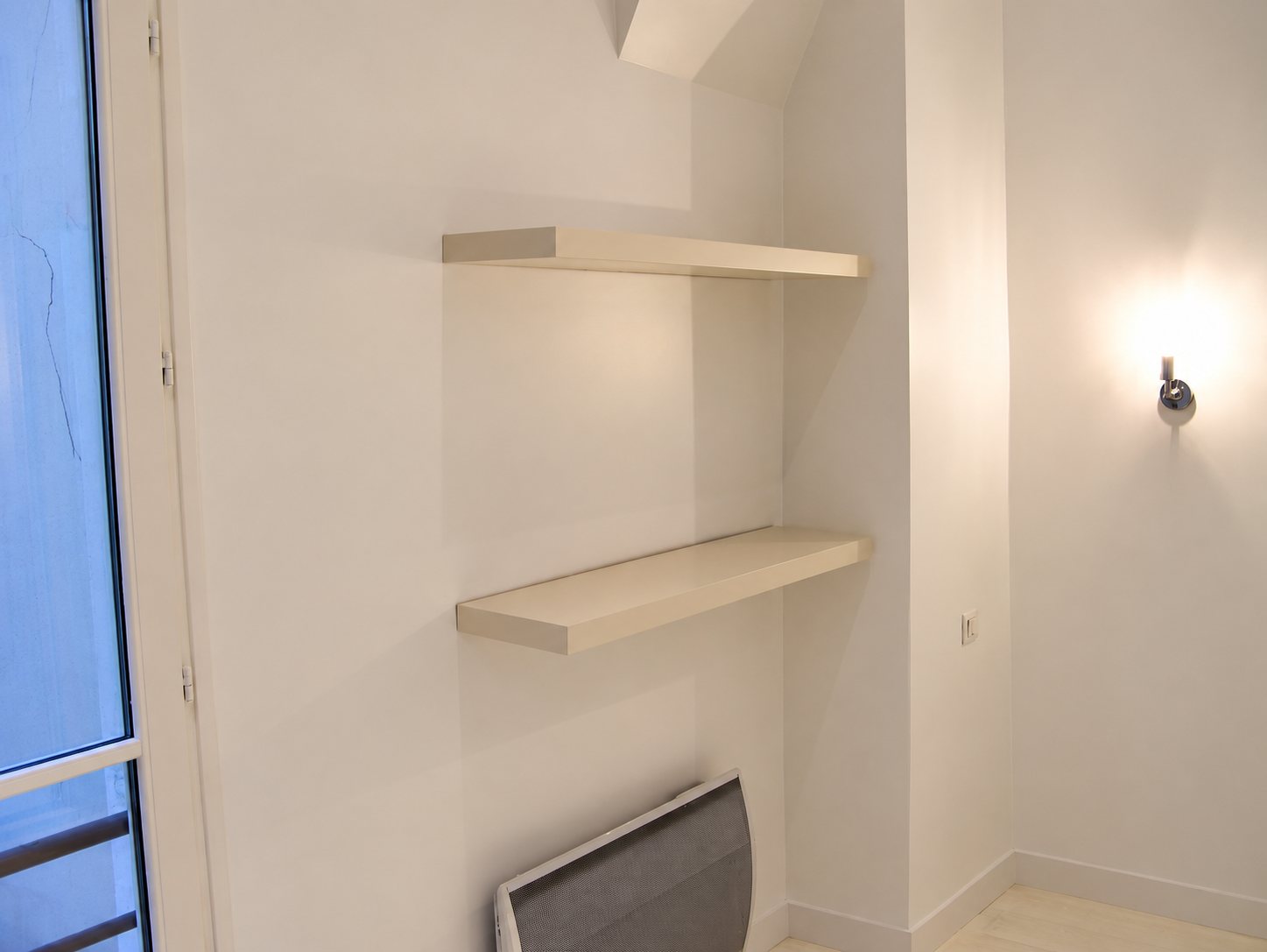 Deux étagères blanches fixées au mur d'une pièce rénovée, avec un radiateur en bas, une applique allumée et une porte-fenêtre à gauche.