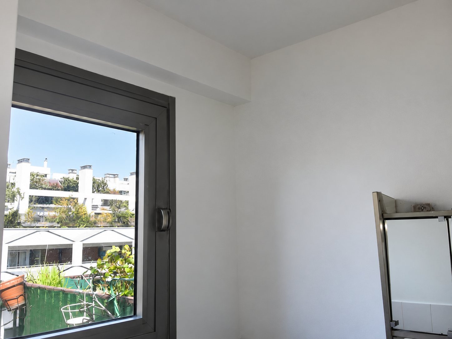 Une fenêtre à côté d'un mur blanc fraîchement peint, avec vue sur un balcon et des immeubles en arrière-plan.