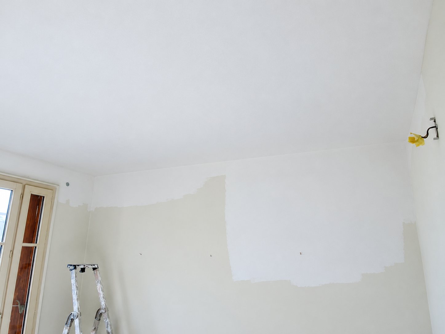 Pièce en cours de rénovation avec un mur et un plafond blancs partiellement repeints, une fenêtre à gauche et un escabeau taché de peinture en bas.