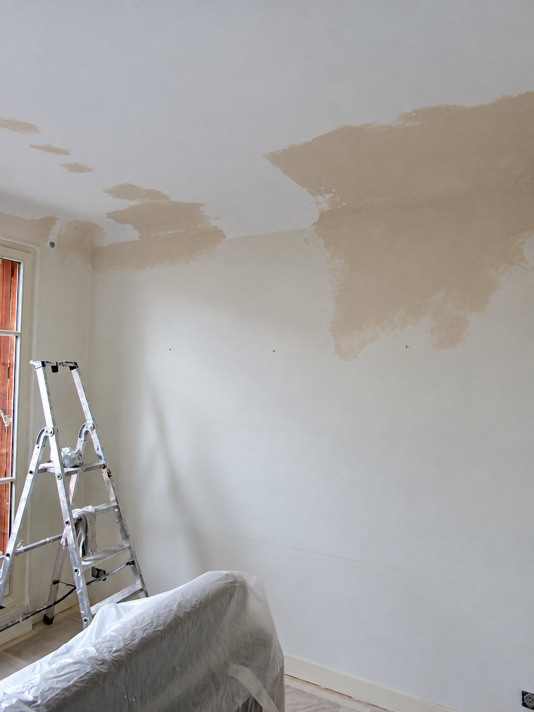 Pièce vide avec un mur blanc et un plafond marqués de grandes taches beige d'humidité, un escabeau en aluminium à gauche et un meuble recouvert d'une bâche en plastique au premier plan.