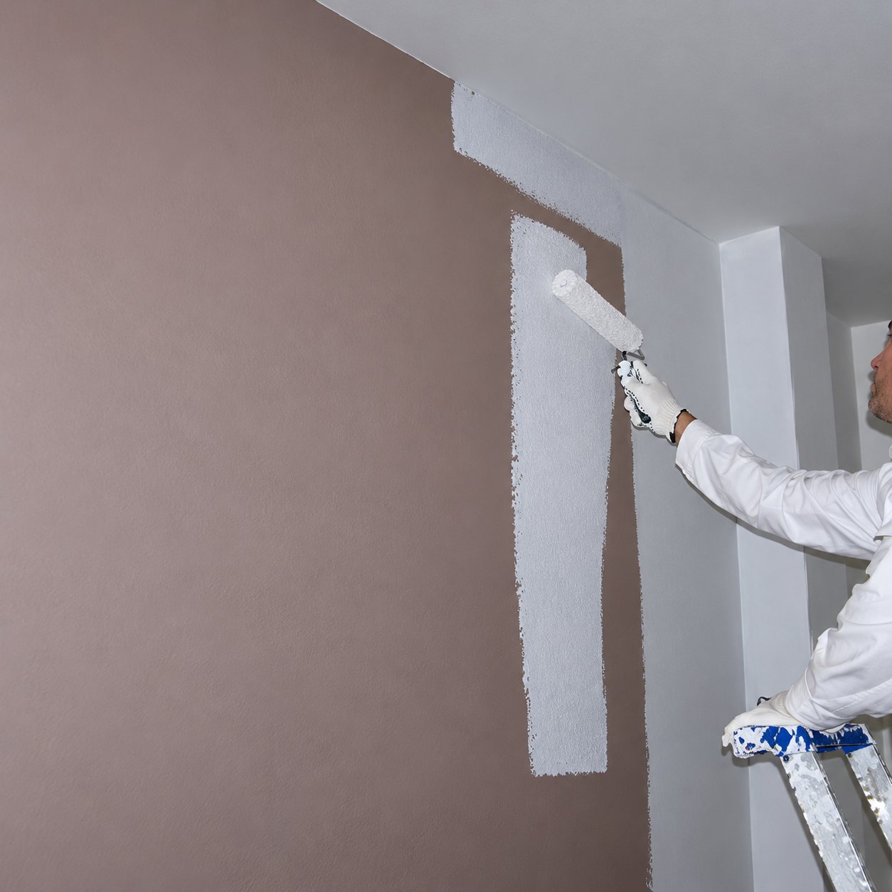 Un peintre en tenue blanche passe un rouleau de peinture blanche sur un mur intérieur d'abord beige rosé.