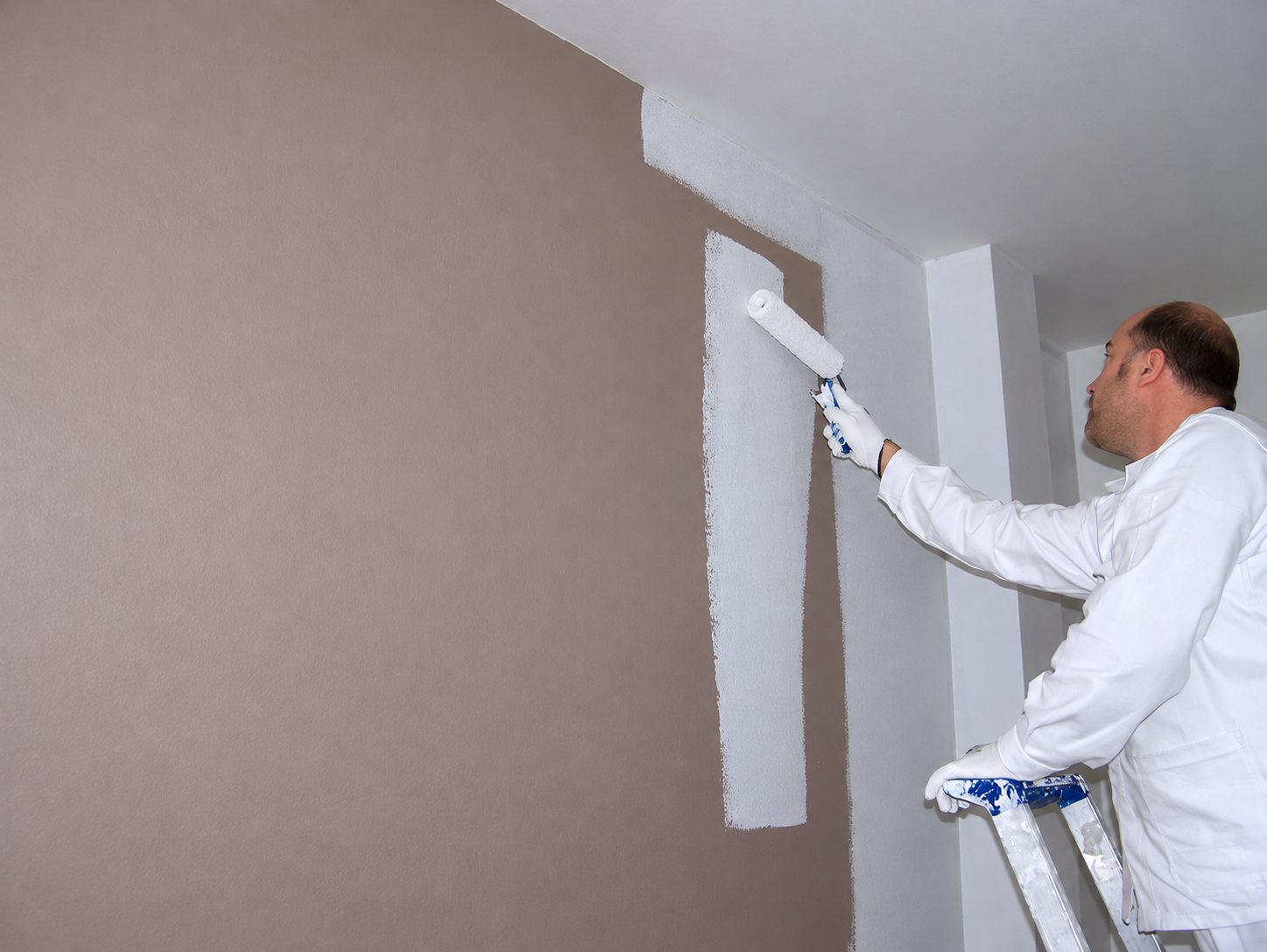 Un peintre en tenue blanche, debout sur un escabeau, passe un rouleau de peinture blanche sur un mur d'abord teinté de taupe.