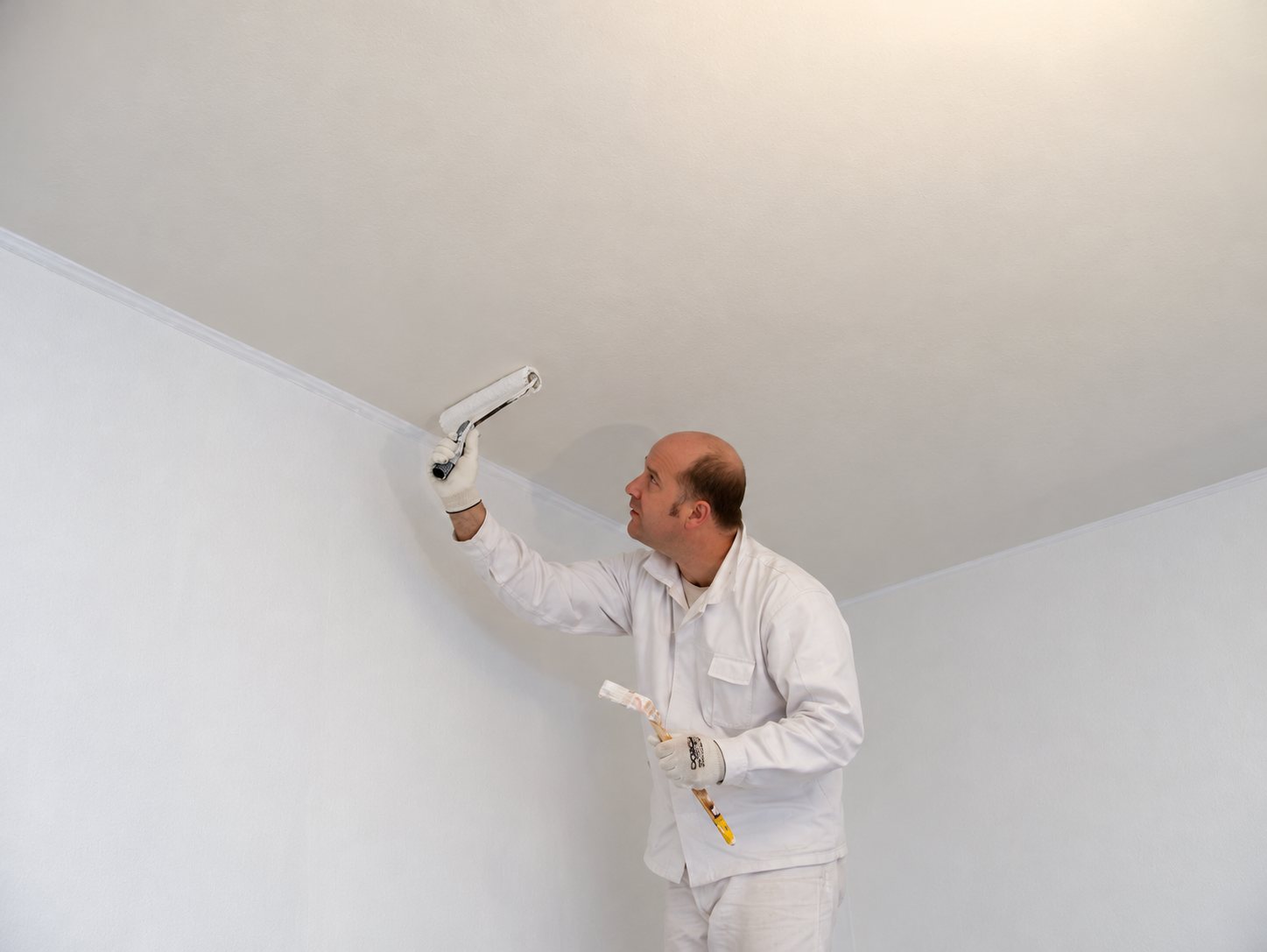 Un peintre en vêtements blancs passe un rouleau de peinture sur le plafond d'une pièce aux murs blancs.