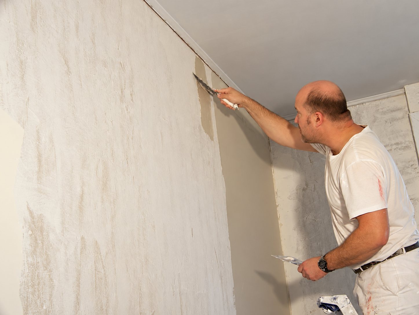 Un homme en tenue blanche applique de l'enduit sur un mur intérieur à l'aide d'un couteau à enduire.
