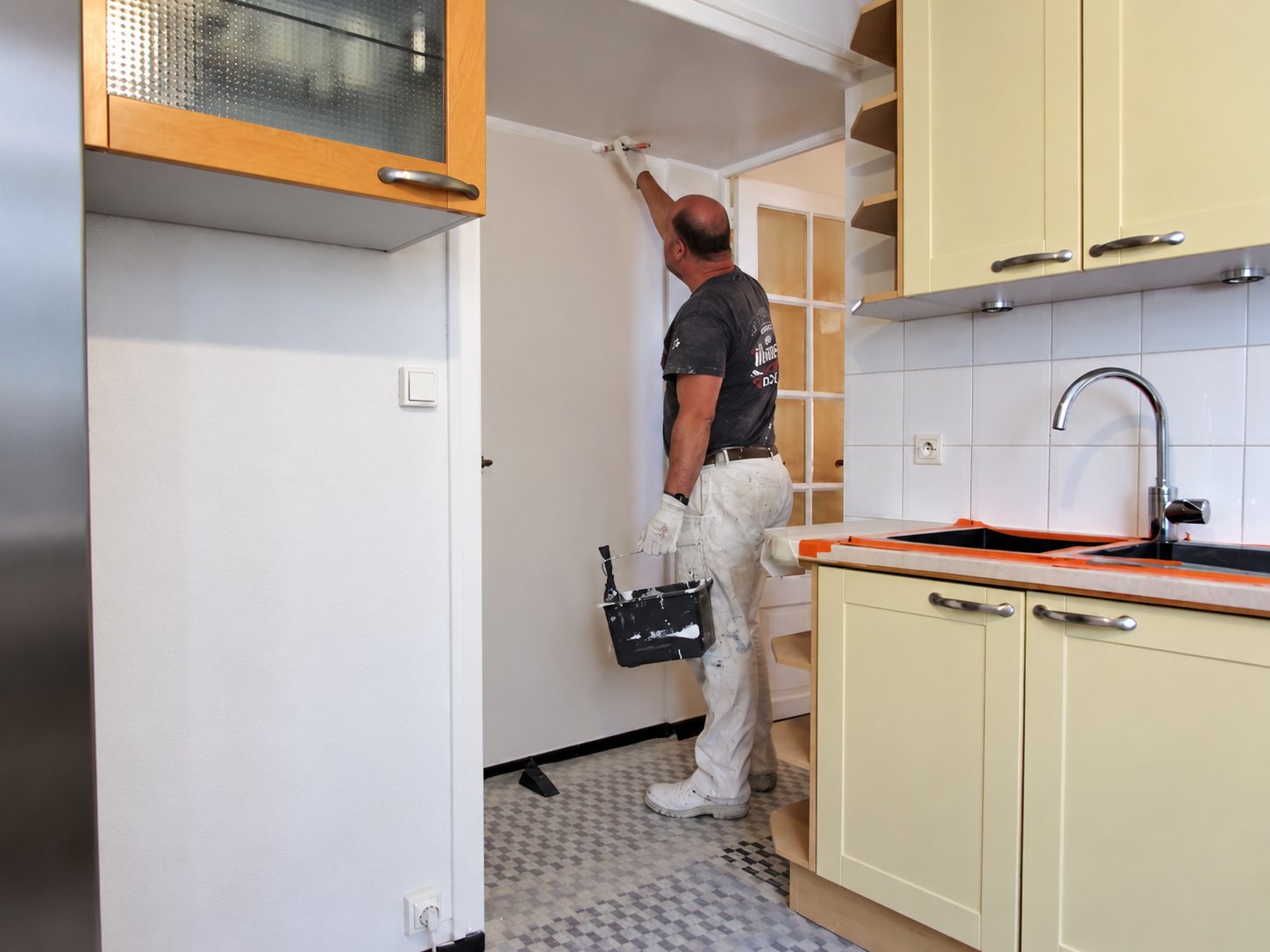 Un peintre en tenue blanche peint le haut d'un mur dans une petite cuisine aux placards beige clair, un seau à la main.