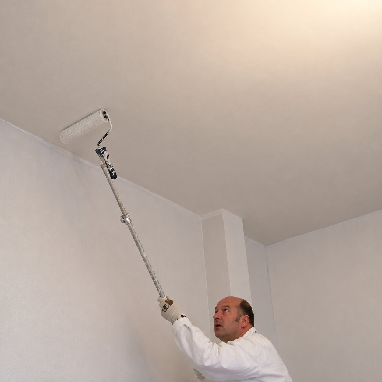 Un peintre en tenue blanche peint le plafond d'une pièce à l'aide d'un rouleau fixé sur une longue perche.