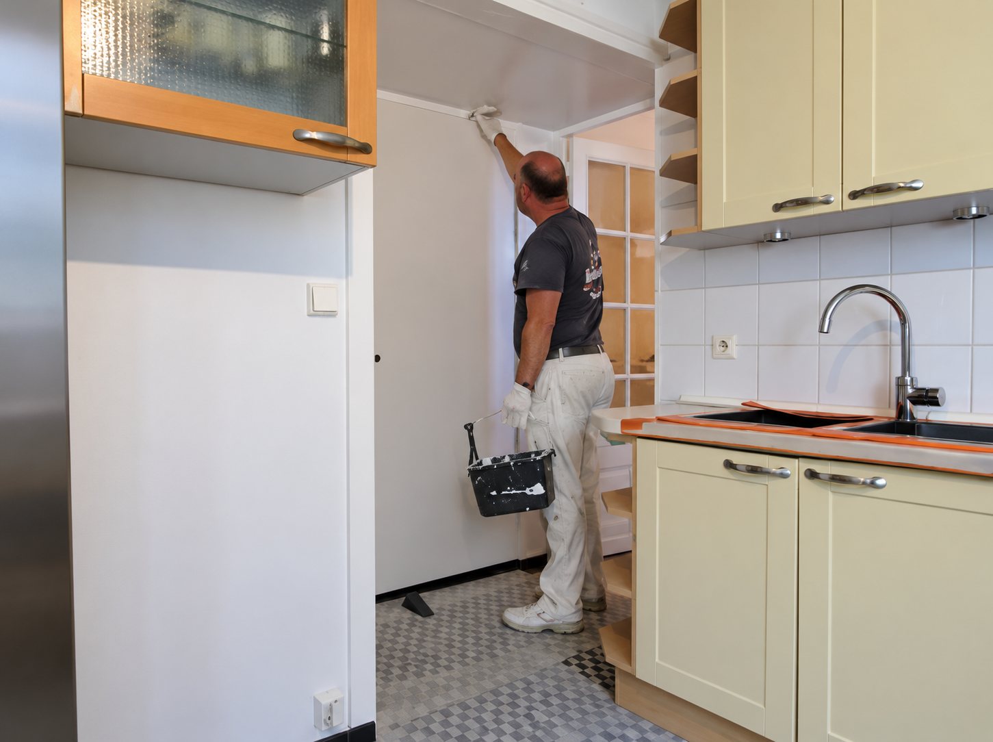 Un peintre en vêtements blancs peint un mur blanc dans une cuisine aux placards beige clair, avec un seau posé au sol.