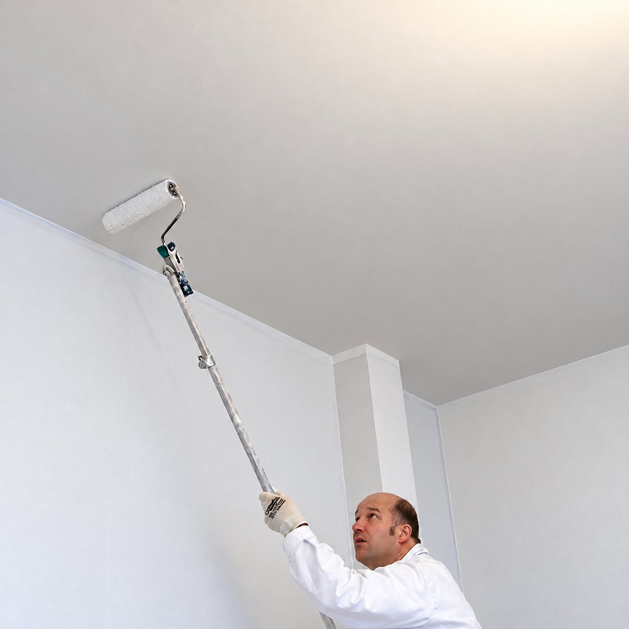 Un peintre en tenue blanche peint le plafond d'une pièce avec un rouleau fixé sur une longue perche.