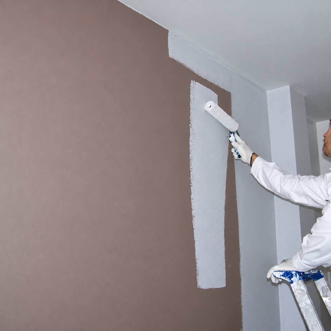 Un peintre en vêtements blancs, debout sur un escabeau, passe un rouleau de peinture blanche sur un mur beige rosé.