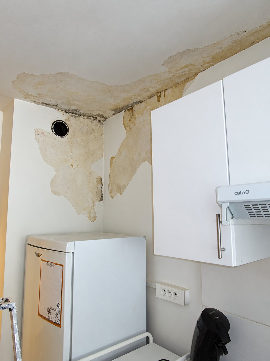 Dans un angle de cuisine, la peinture du plafond et du mur s'écaille à cause de l'humidité, au-dessus d'un petit réfrigérateur et d'un placard blanc équipé d'une hotte.