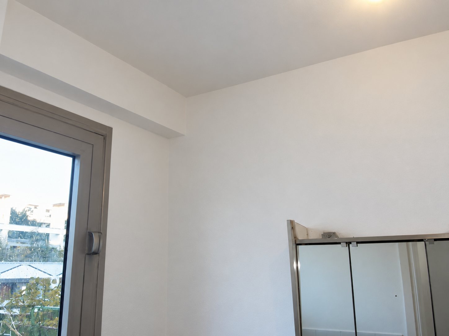 Angle d'une pièce aux murs et plafond fraîchement peints en blanc, avec une fenêtre à gauche et une cabine de douche vitrée en bas à droite.