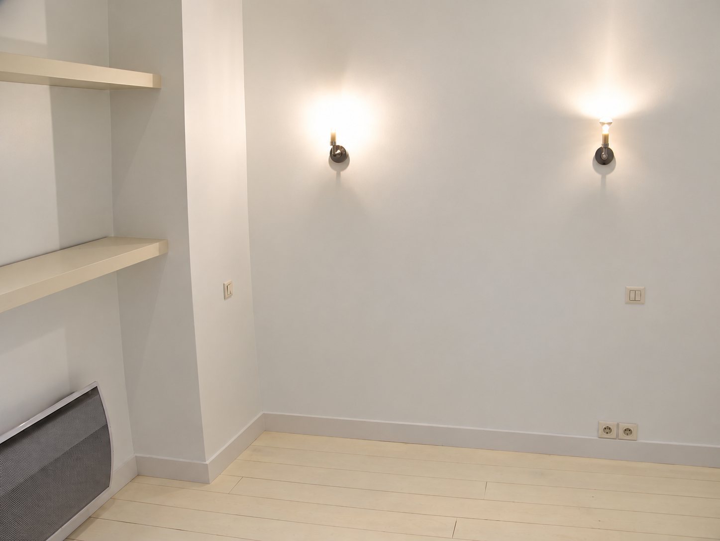 Pièce vide fraîchement rénovée aux murs et au parquet blancs, éclairée par deux appliques murales, avec un radiateur gris et des étagères intégrées.