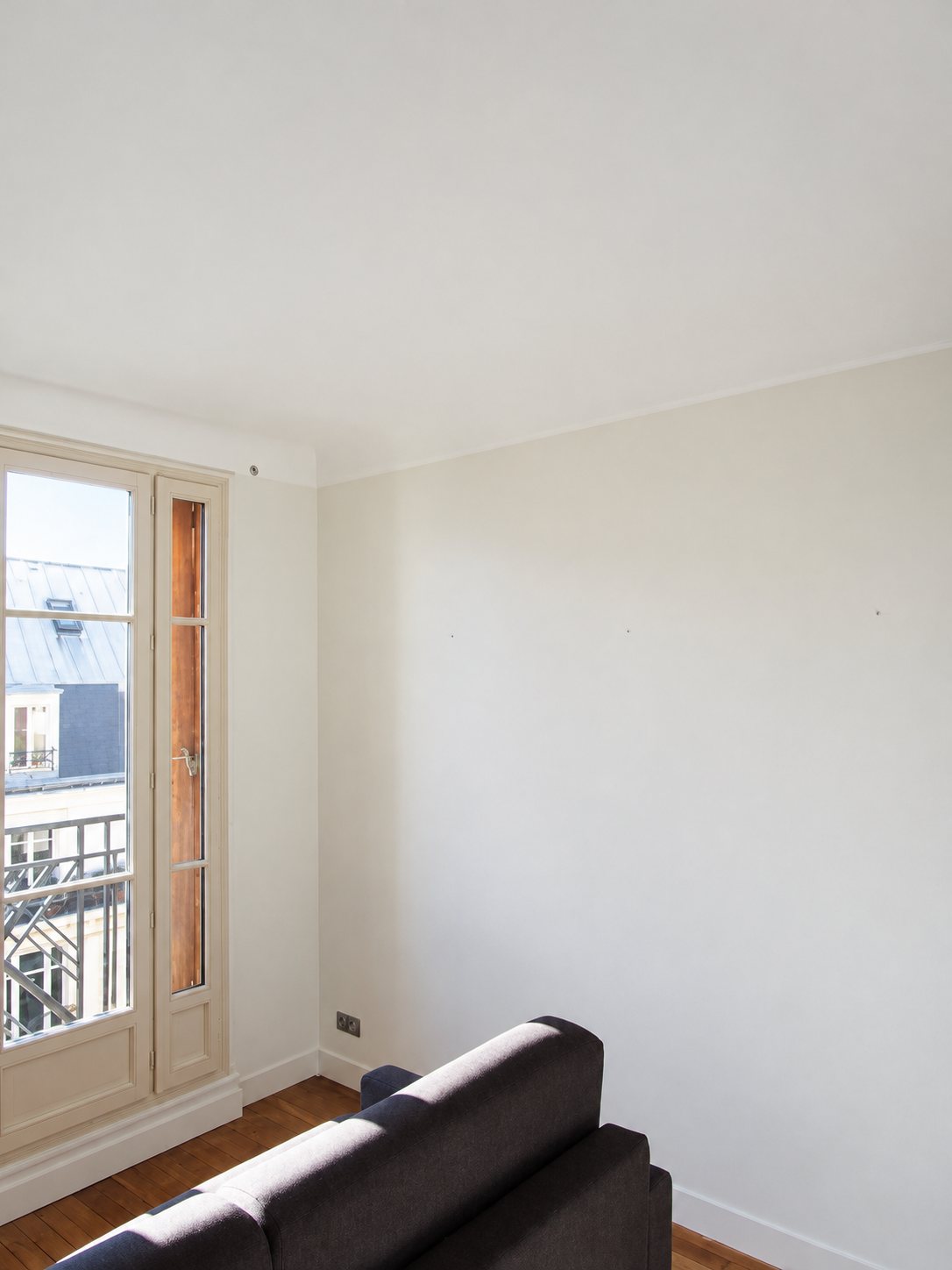 Pièce fraîchement repeinte en blanc avec une porte-fenêtre donnant sur un balcon, un parquet en bois et un canapé sombre au premier plan.