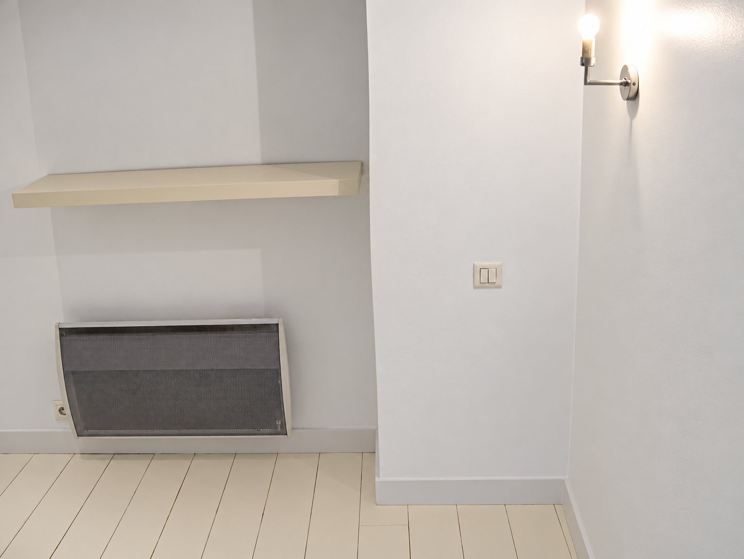 Intérieur rénové aux murs et au parquet peints en blanc, avec une étagère, un radiateur électrique gris, un interrupteur et une applique allumée.