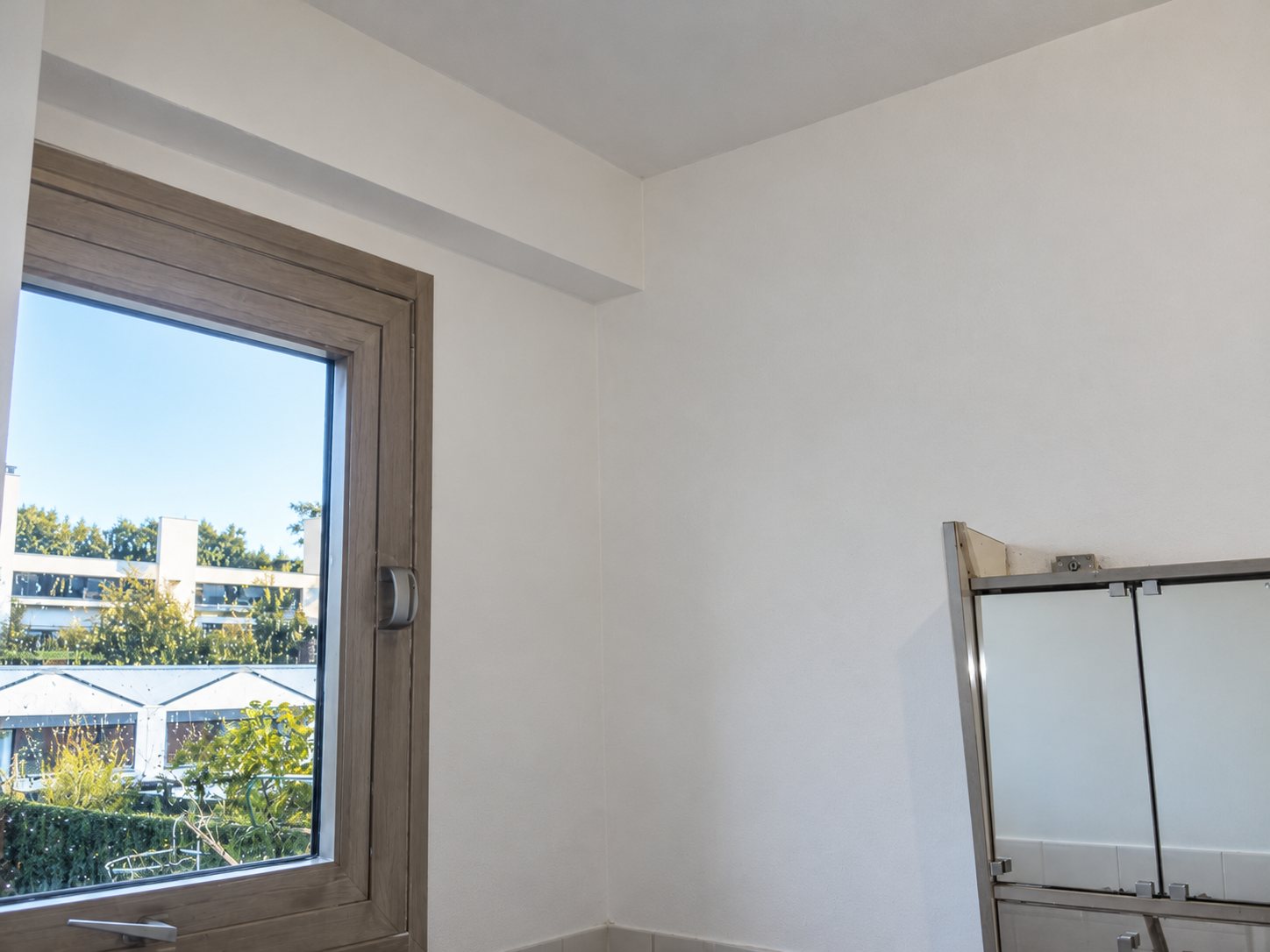 Angle d'une pièce aux murs et au plafond fraîchement peints en blanc, avec une fenêtre donnant sur des immeubles et un cadre métallique de douche à droite.