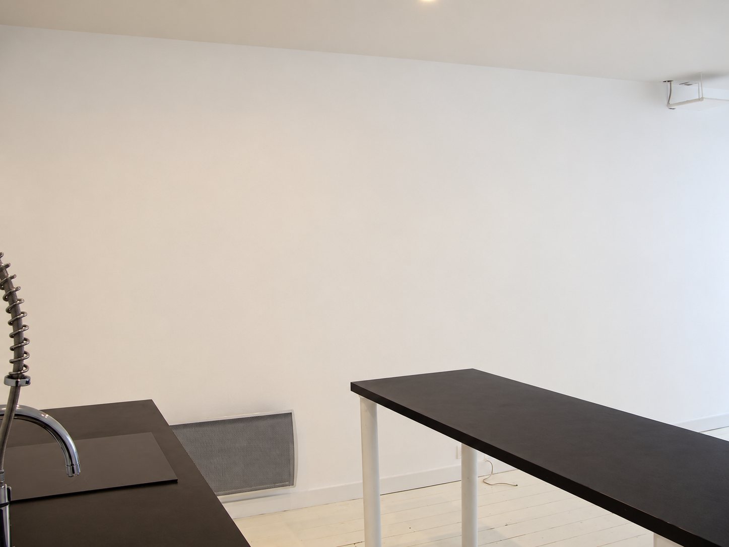 Intérieur d'une pièce vide aux murs blancs avec un évier à douchette, un plan de travail noir, une table haute noire et un radiateur gris.