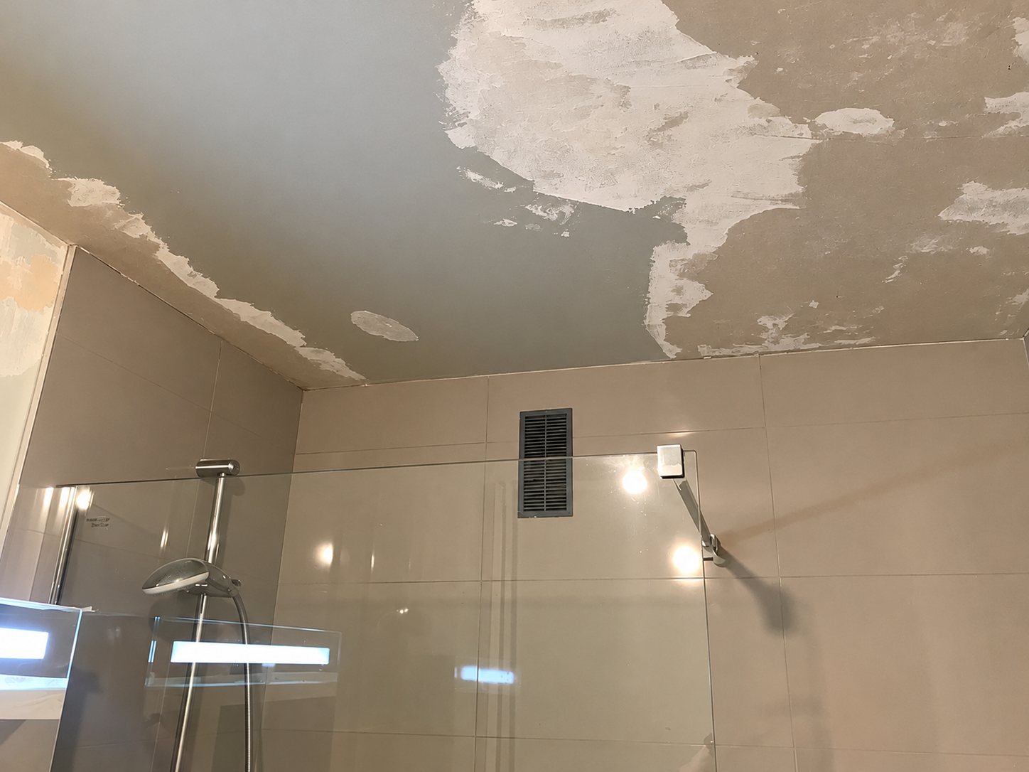 Plafond de salle de bain dont la peinture s'écaille par plaques, au-dessus d'une douche à paroi de verre et d'une grille de ventilation.