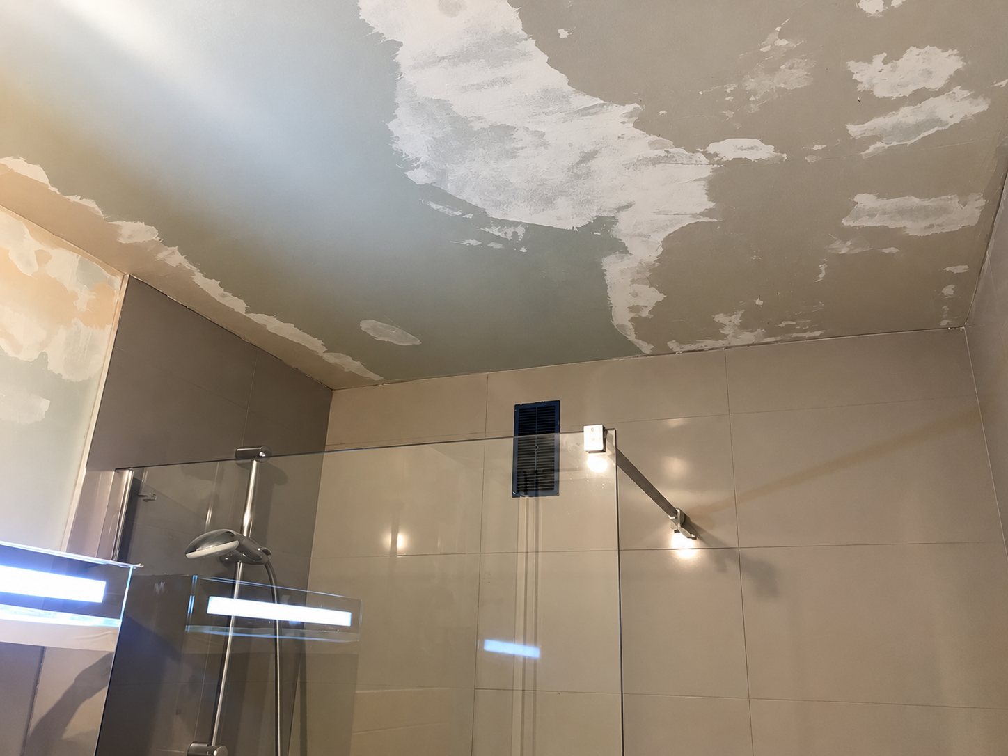 Plafond de salle de bain dont la peinture s'écaille et présentant des zones d'enduit, au-dessus d'une douche à l'italienne avec paroi en verre.