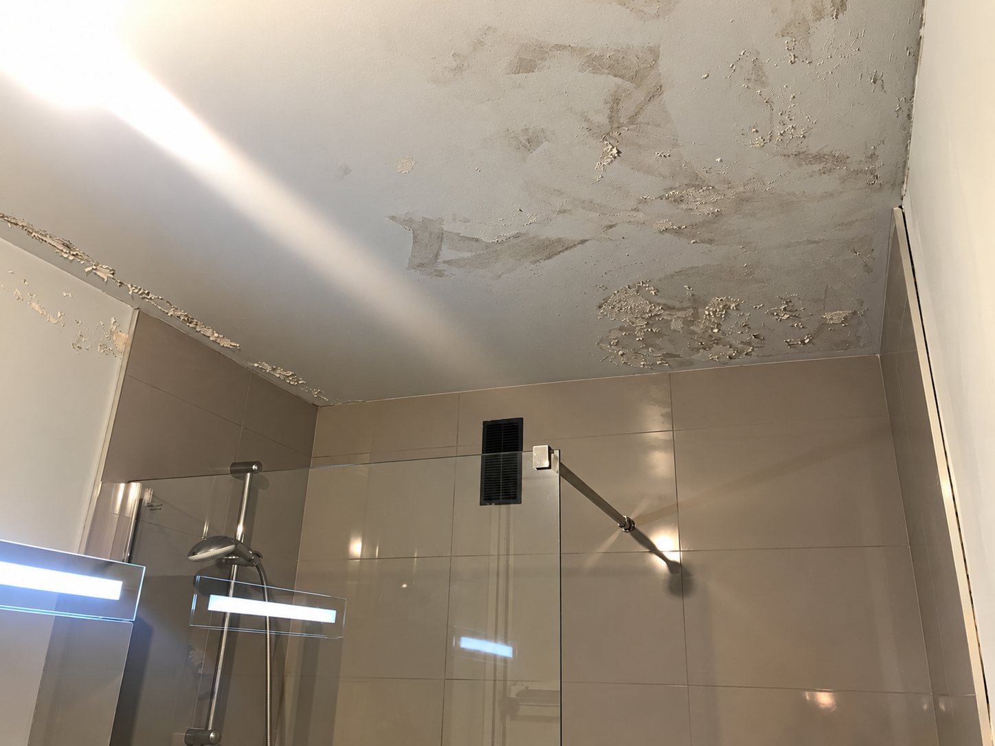 Plafond de salle de bain dont la peinture s'écaille et présente des taches d'humidité au-dessus d'une douche vitrée carrelée.