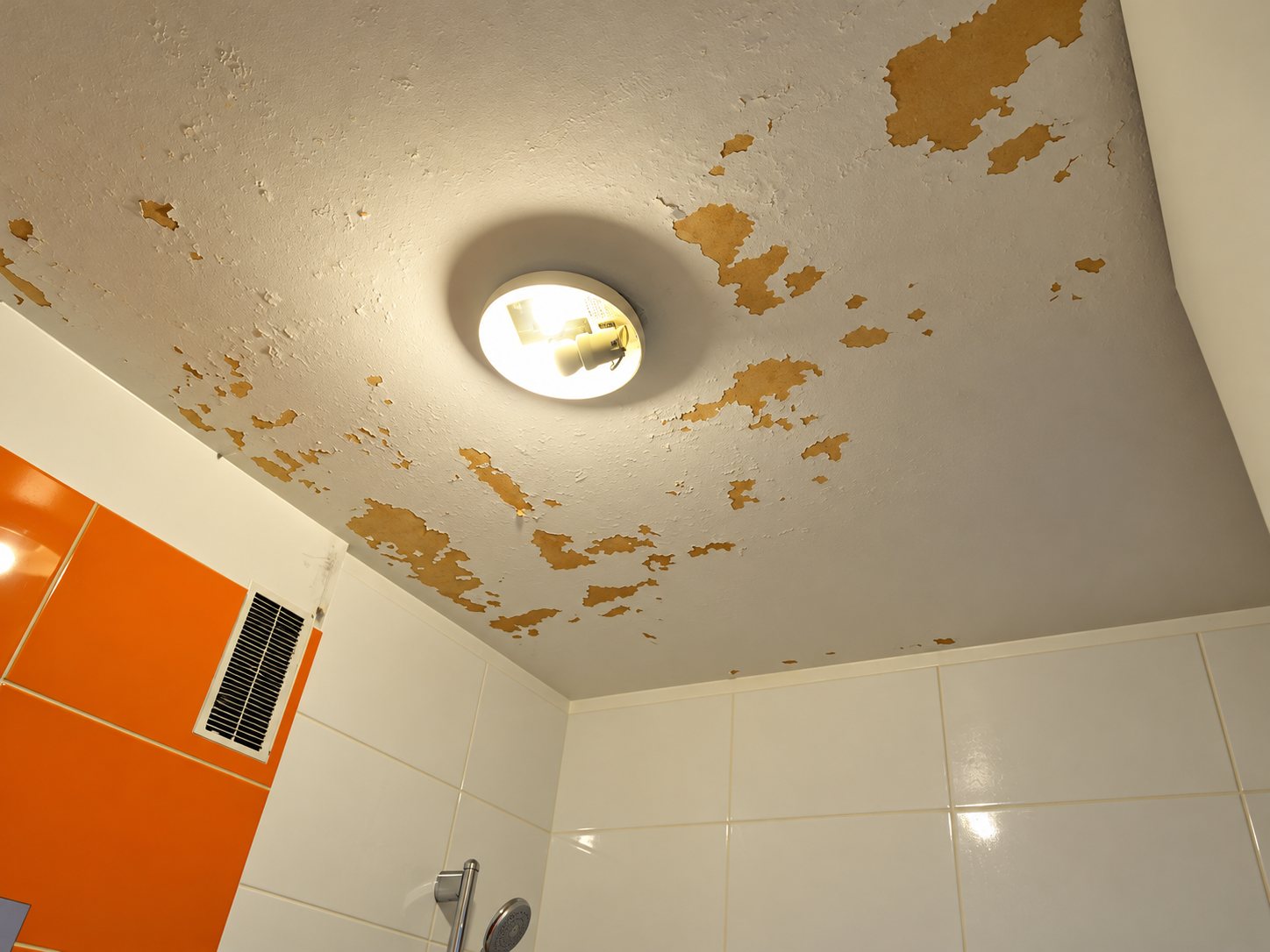 Plafond de salle de bain dont la peinture s'écaille par plaques, avec un plafonnier allumé, des carreaux blancs et orange et une grille d'aération.