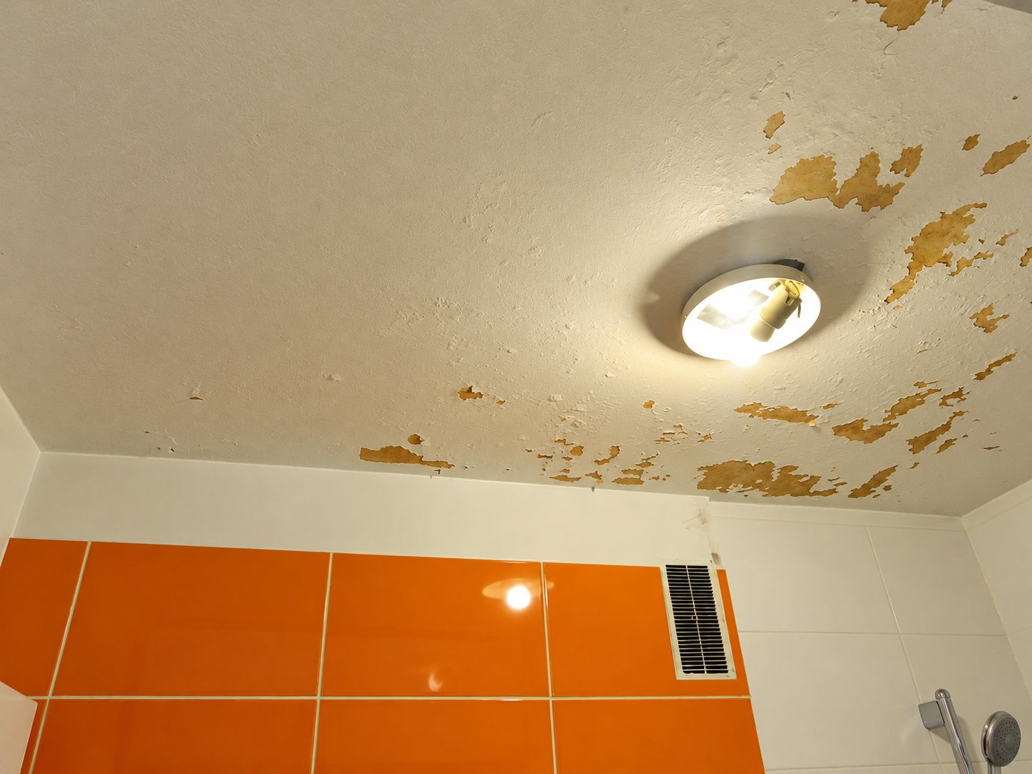 Plafond de salle de bain dont la peinture s'écaille par plaques, avec un plafonnier allumé, une grille d'aération et un carrelage orange au mur.