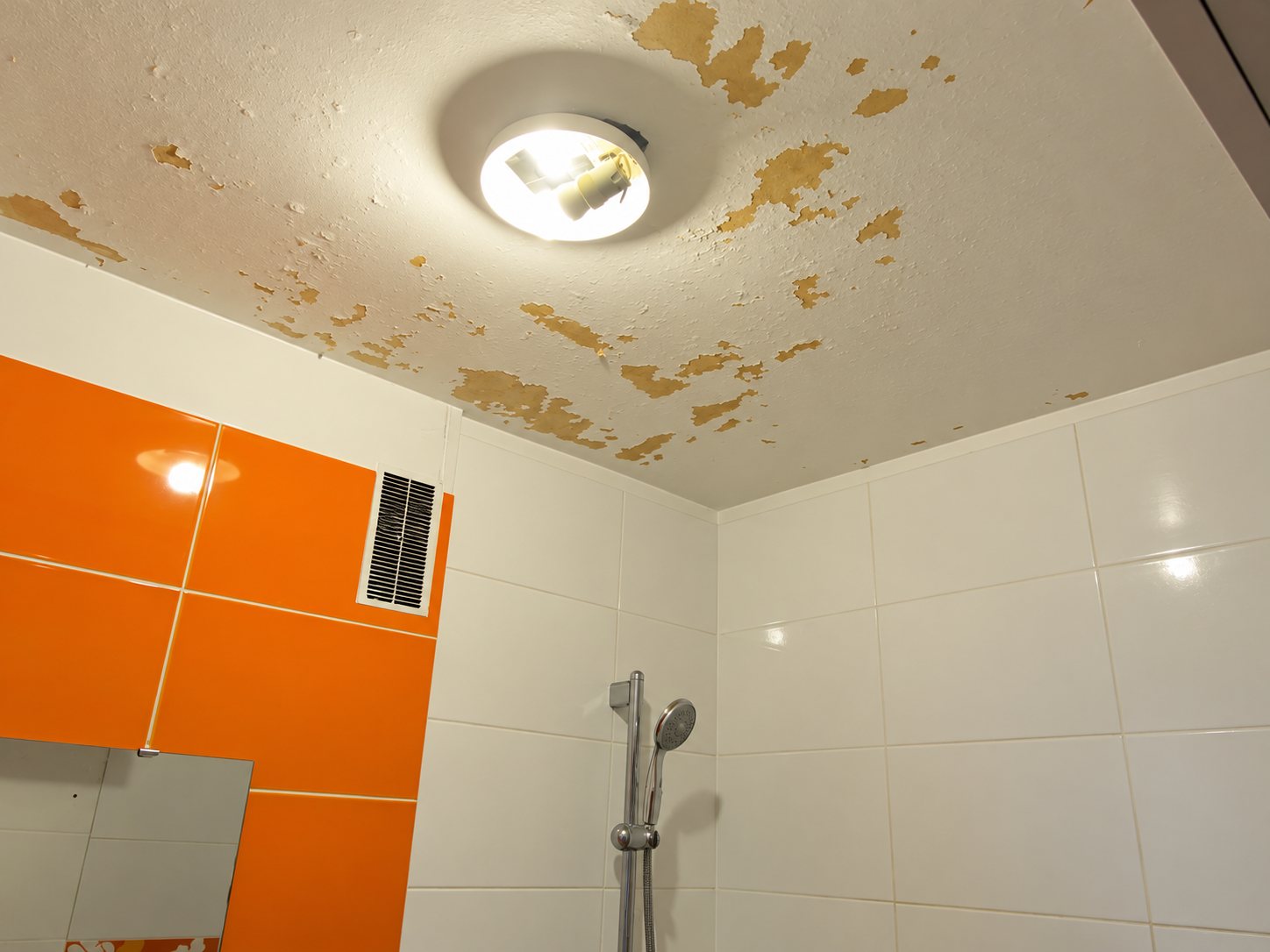 Plafond d'une salle de bains dont la peinture s'écaille autour d'un plafonnier allumé, avec carrelage orange et blanc et une douchette au mur.