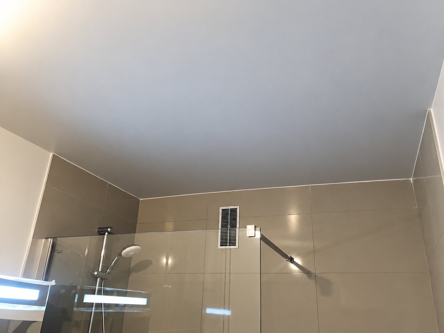 Plafond lisse de couleur taupe au-dessus d'une douche à l'italienne avec paroi en verre, carrelage beige et grille de ventilation.
