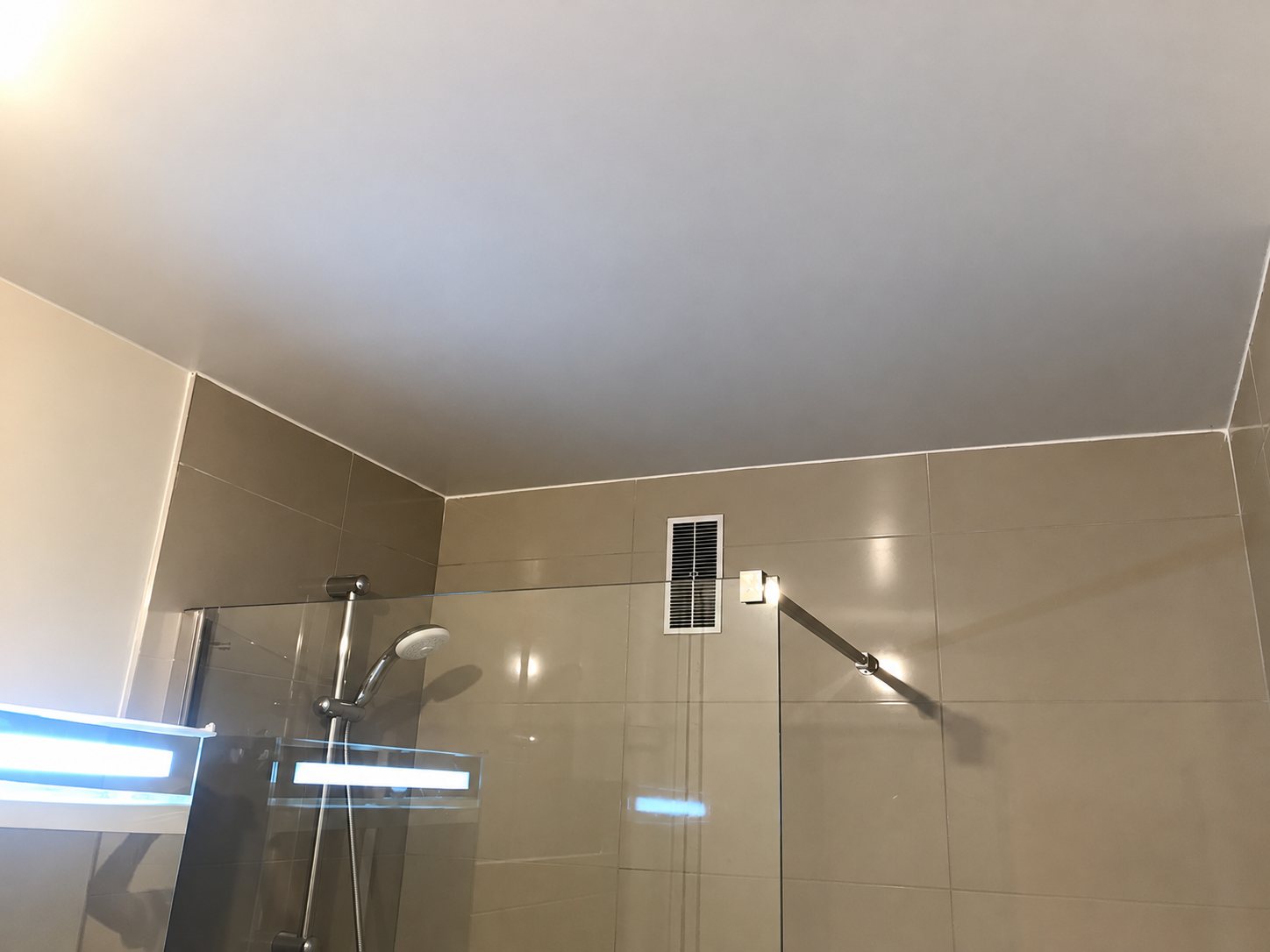 Plafond tendu beige et lisse au-dessus d'une douche à l'italienne avec paroi en verre, carrelage beige et grille de ventilation.