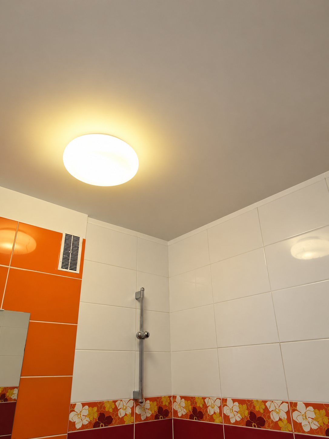 Un plafonnier rond lumineux au plafond d'une salle de bain aux murs carrelés de blanc et d'orange, avec une barre de douche et une frise fleurie.