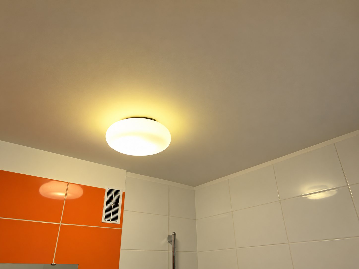Un plafonnier rond en verre opalin éclaire le plafond d'une salle de bains aux murs carrelés de blanc et d'orange, avec une grille d'aération.