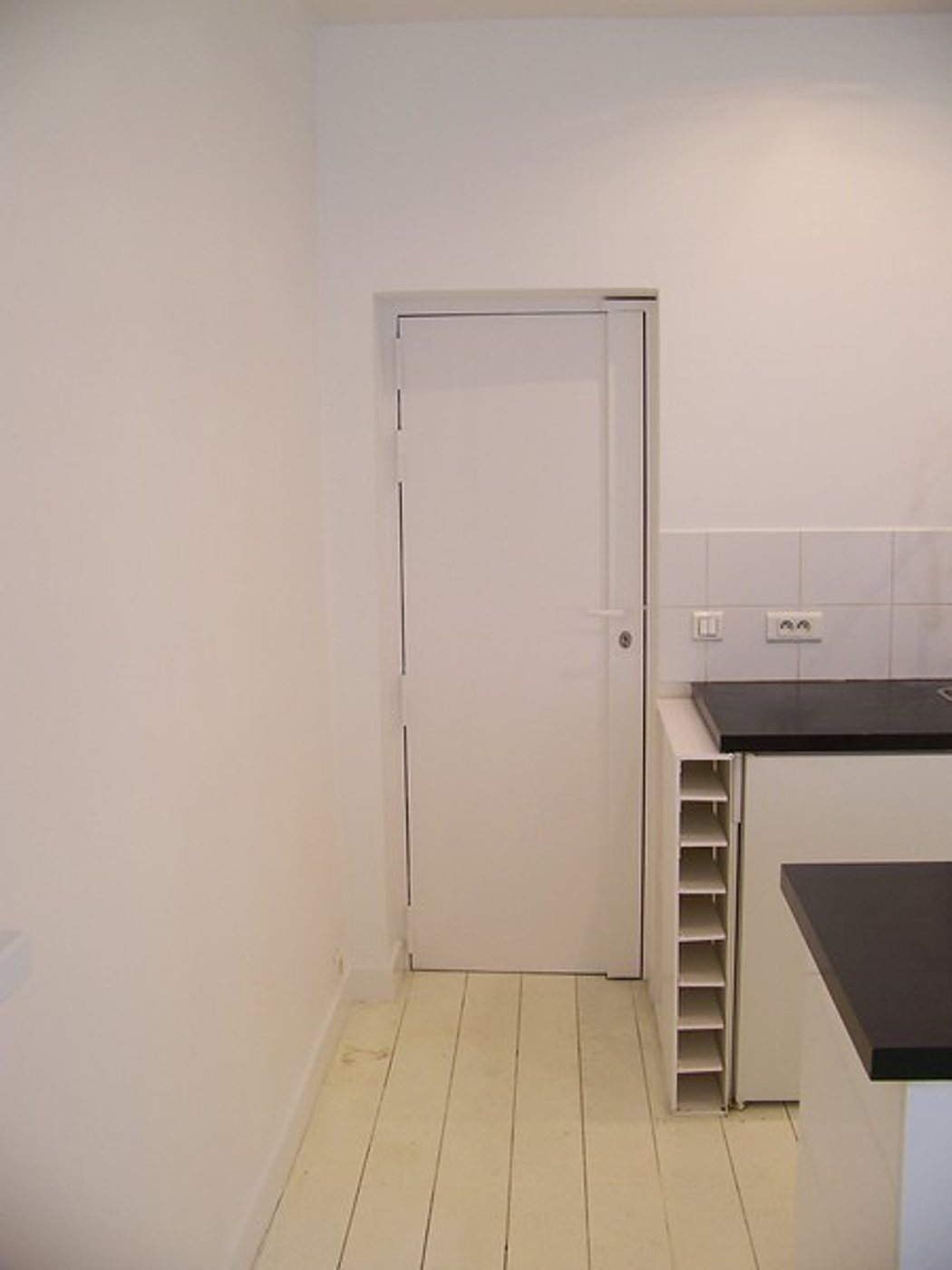 Une porte blanche encadrée dans un mur clair, à côté d'un meuble casier à bouteilles et d'un plan de travail noir, sur un plancher en lattes peintes en blanc.