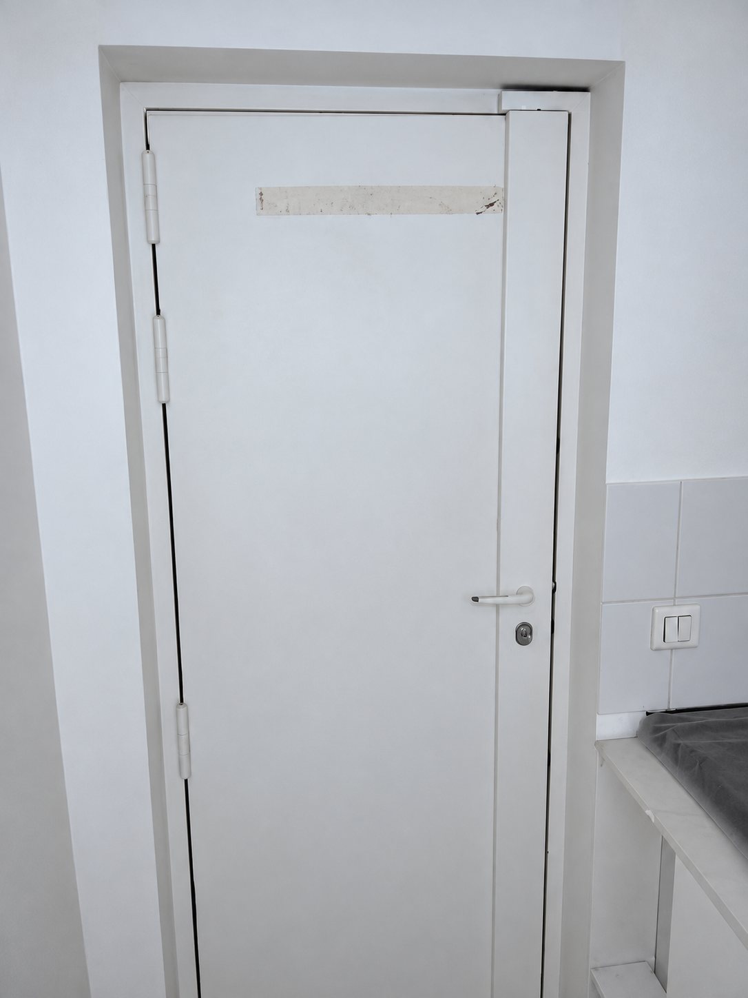 Une porte blanche fermée avec poignée et serrure, encadrée par des murs blancs, à côté d'un plan de travail et d'une prise électrique.