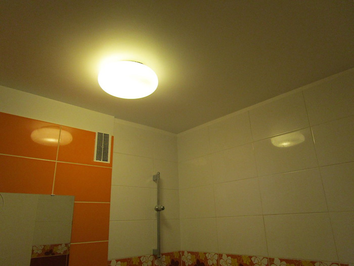 Salle de bain rénovée avec carrelage mural blanc et orange à motifs de fleurs, grille de ventilation, barre de douche et plafonnier rond allumé.