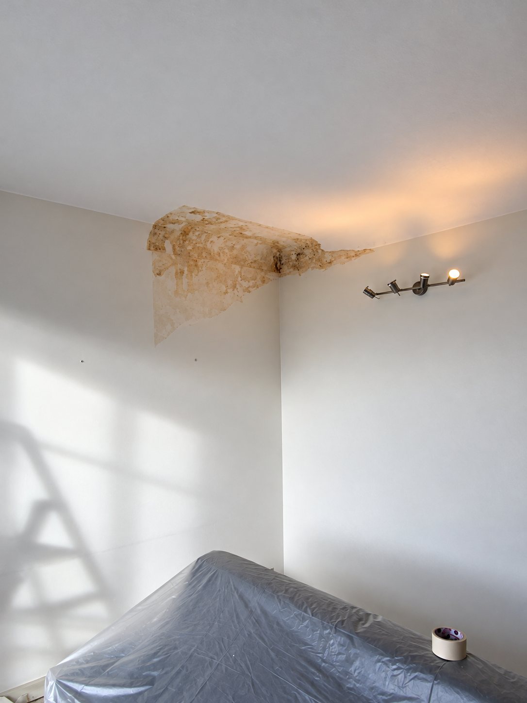 Dans une pièce aux murs blancs, une grande tache d'humidité jaunâtre marque l'angle du plafond, au-dessus d'un meuble recouvert d'une bâche et d'un rouleau de ruban adhésif.