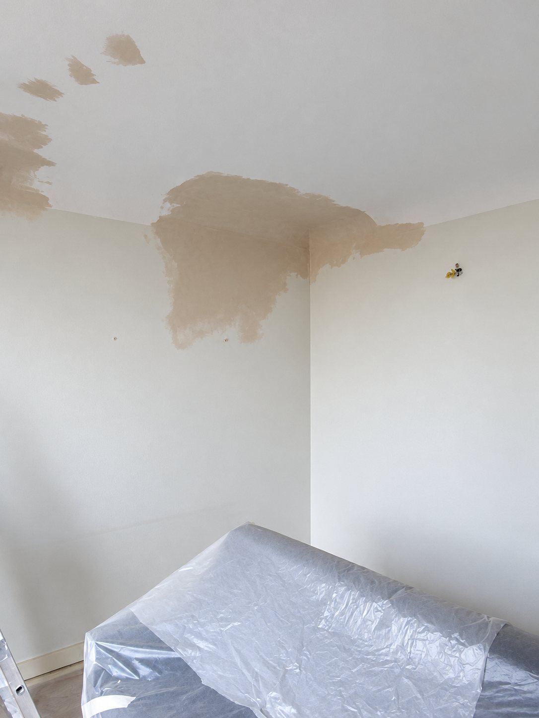 Angle d'une pièce avec une grande tache d'humidité brune au plafond et au mur, et un meuble recouvert d'une bâche plastique au premier plan.